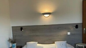 A neatly made bed with white linens and pillows, a bedside table with a plant, and a wall-mounted light fixture. - Duplex 6 people in Corsica in residence with swimming pool (Serra-di-Ferro)