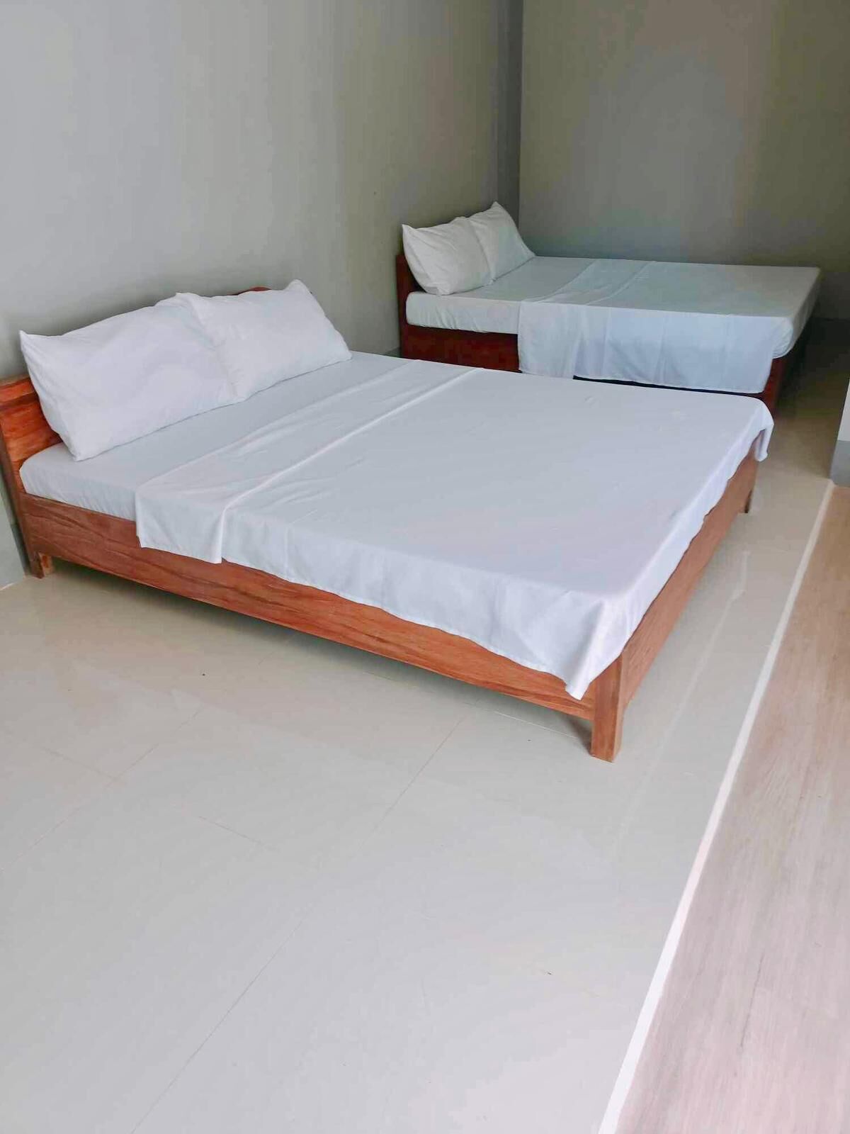 Dos camas de madera con ropa de cama blanca en una habitación con paredes lisas y piso de baldosas.
