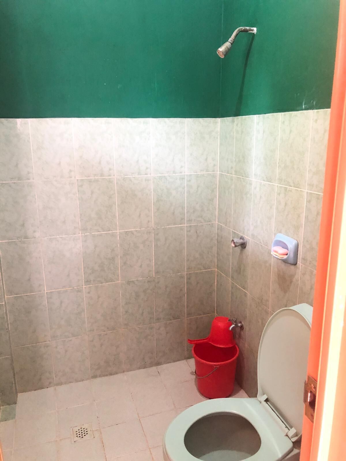 A small, tiled bathroom with a toilet, a red bucket, and a showerhead.