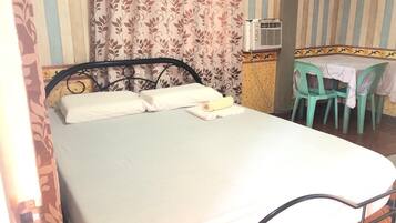 A single bed with white linens, a metal frame, and a small table with a white tablecloth and two green chairs.
