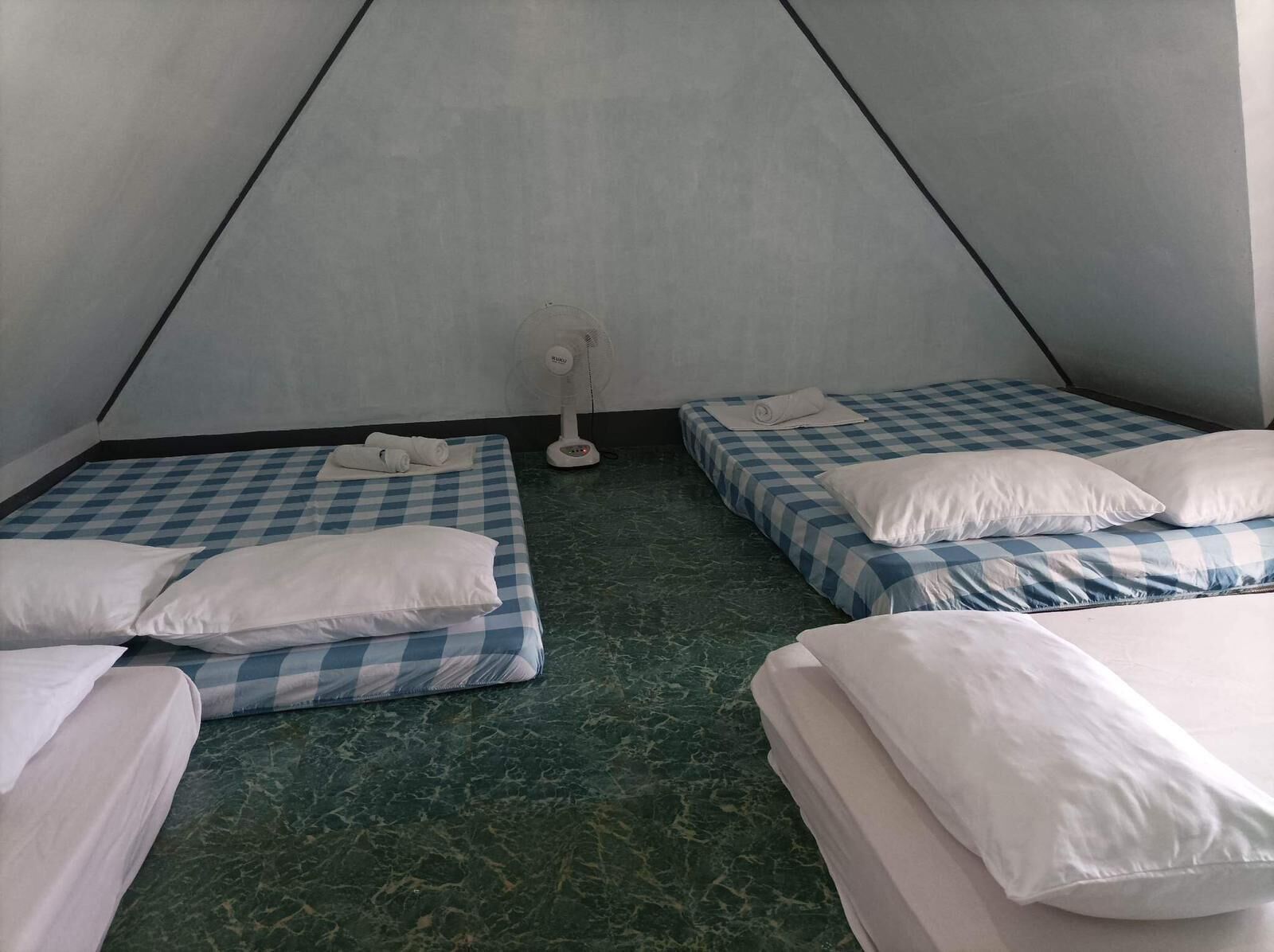 Une chambre avec deux lits, chacun recouvert d’une couette à carreaux bleus et blancs, et un ventilateur placé au centre de la pièce.