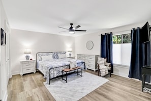 Ein Schlafzimmer mit Bett, Nachttischen, einem Schreibtisch, einem Stuhl, einem Deckenventilator und einem Fenster mit Vorhängen.