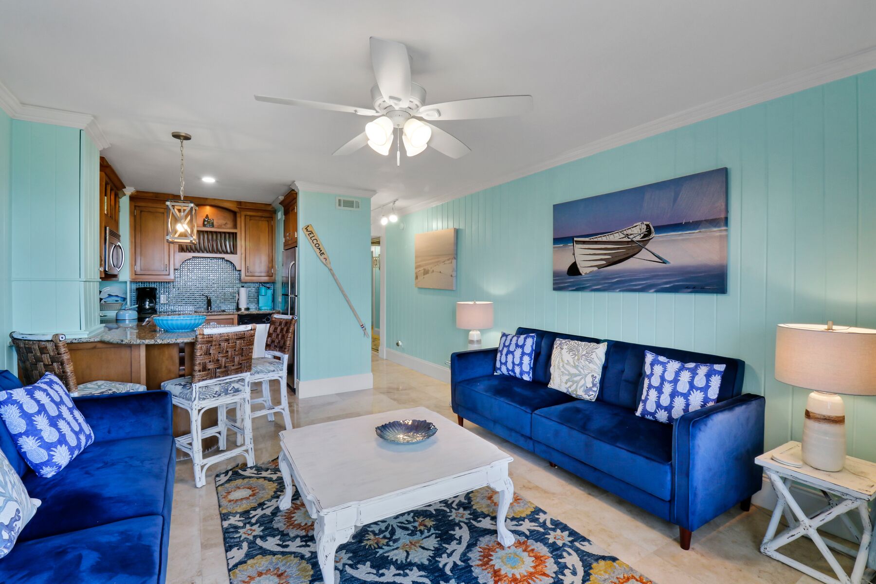 A living room with a blue sofa, white coffee table, and a patterned rug. There is a kitchen area with wooden cabinets and a hanging light fixture.