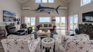 A spacious living room with a high ceiling, a large mounted shark artwork, a television, and a view of the outdoors through large sliding doors. - Rum & Coconuts-You'll be livin' the Caribbean dream! (Hope Town, Bahamas)