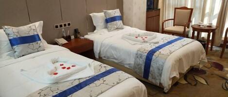 A hotel room with two beds, each with a blue-striped bedspread and matching pillows. There is a small wooden nightstand with a lamp and a telephone. A chair and a small table are visible in the corner.