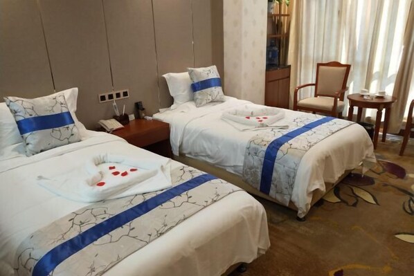 A hotel room with two beds, each with a blue-striped bedspread and matching pillows. There is a small wooden nightstand with a lamp and a telephone. A chair and a small table are visible in the corner.