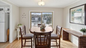 A dining area with a round table, four chairs, a ceiling light, a large window with a view of a lake, and a framed picture on the wall.