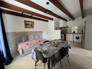 A room with a bed, a dining table with chairs, a kitchen area with a washing machine, and a ceiling fan.