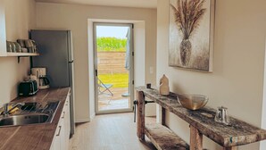 Una cucina moderna con piano di lavoro in legno, forno a incasso e macchina da caffè. Si nota una panca in legno e un tavolo con un vaso e una caffettiera. Una porta dà accesso a un'area esterna con giardino.