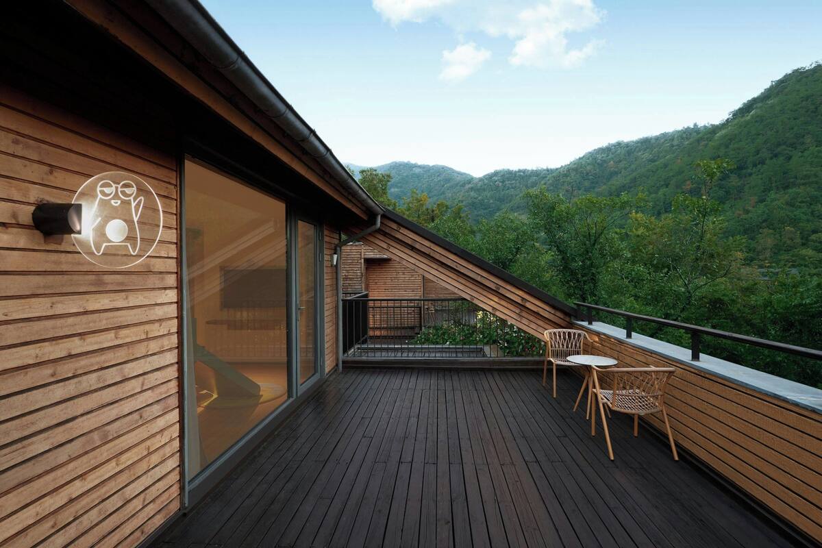 A wooden deck with a glass wall, a small table, and chairs, overlooking a forested hillside.