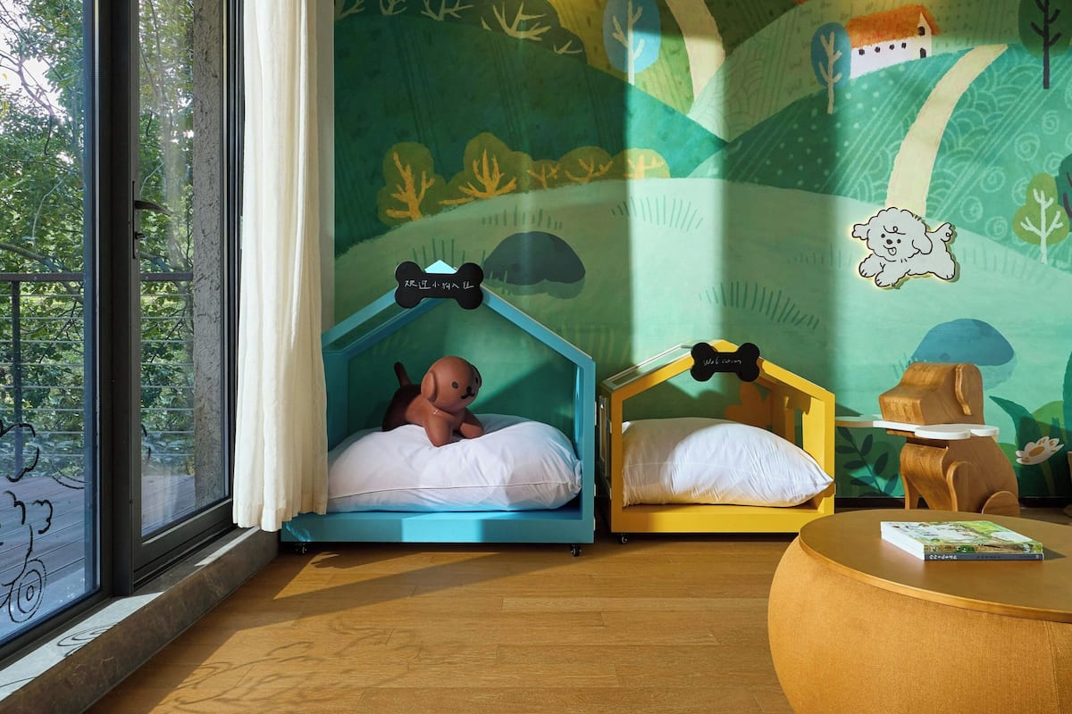 A room with two beds, one with a blue canopy and the other with a yellow one. There is a window with a curtain, a round wooden table, and a wall with a nature-themed mural.