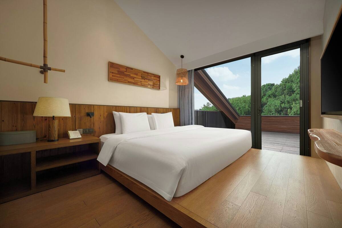 A modern bedroom with a large bed, wooden headboard, a bedside table with a lamp, and a view of a balcony.