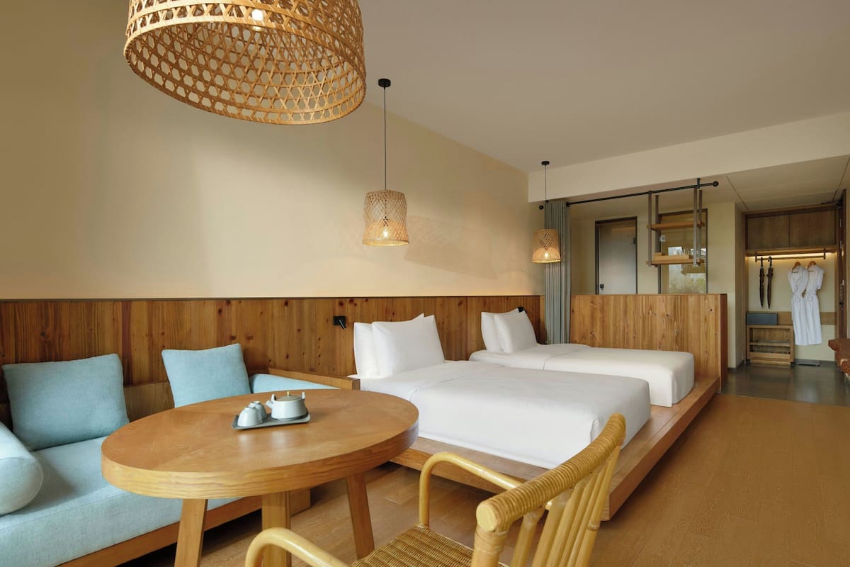 A hotel room with a wooden wall panel, two beds, a round wooden table, and wicker chairs.