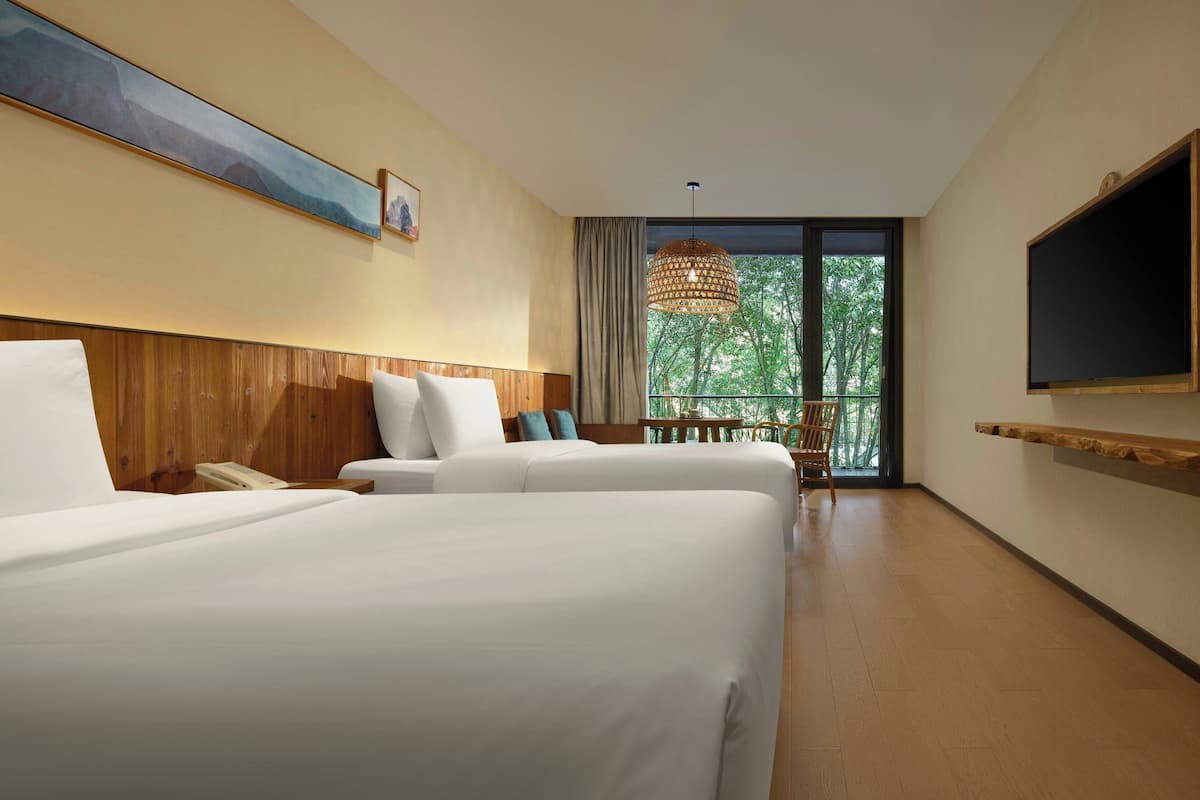 A hotel room with two beds, a wooden headboard, a mounted TV, and a large window with a view of trees.
