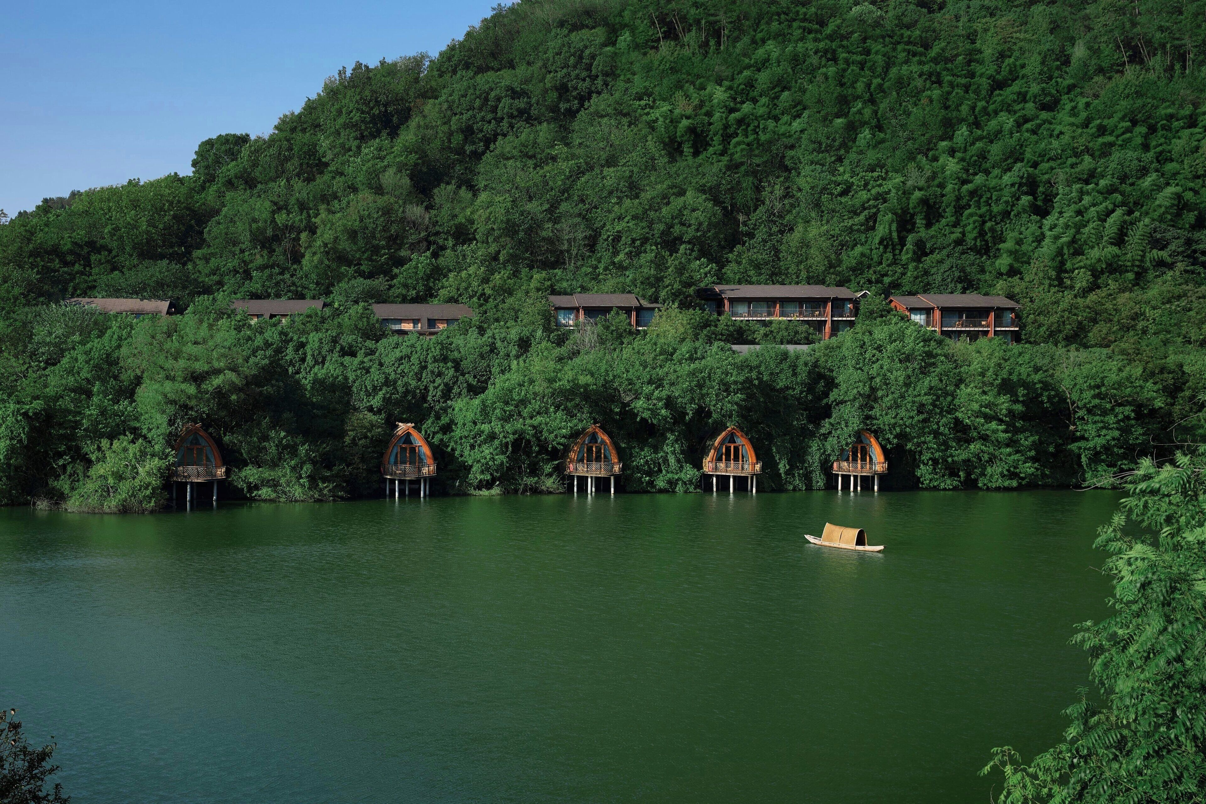 Una hilera de chalets de madera con techos empinados, construidos sobre pilotes sobre un lago tranquilo, rodeados de un bosque denso.