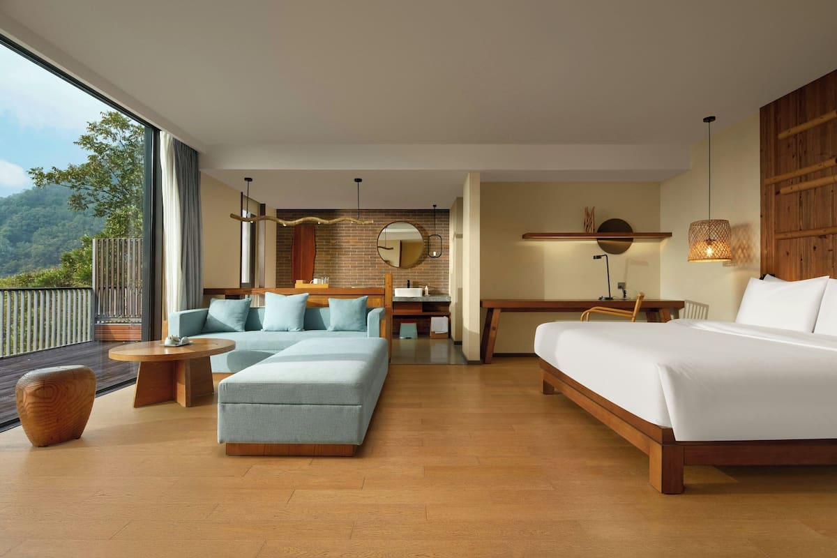 A modern hotel room with a large bed, a sofa, a dining table, and a view of the outdoors.