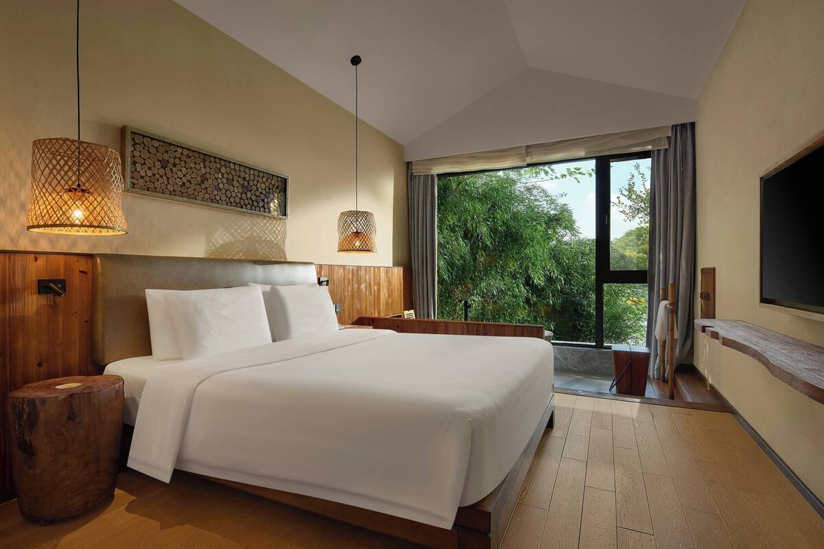 A modern hotel room with a large bed, wooden headboard, and a large window offering a view of greenery.
