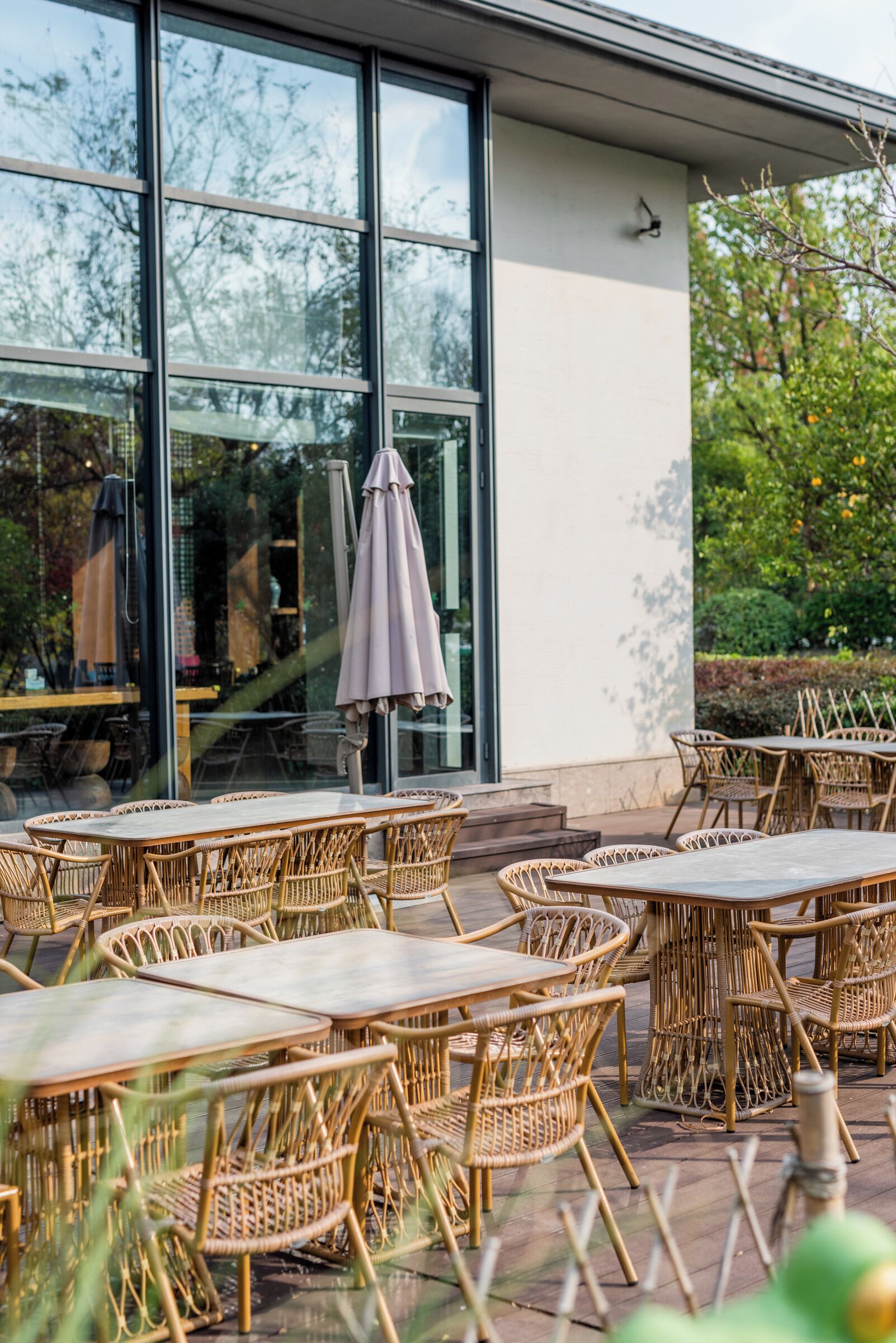 Un espace extérieur aménagé avec des tables et des chaises en osier, de grandes fenêtres et une vue sur les arbres.
