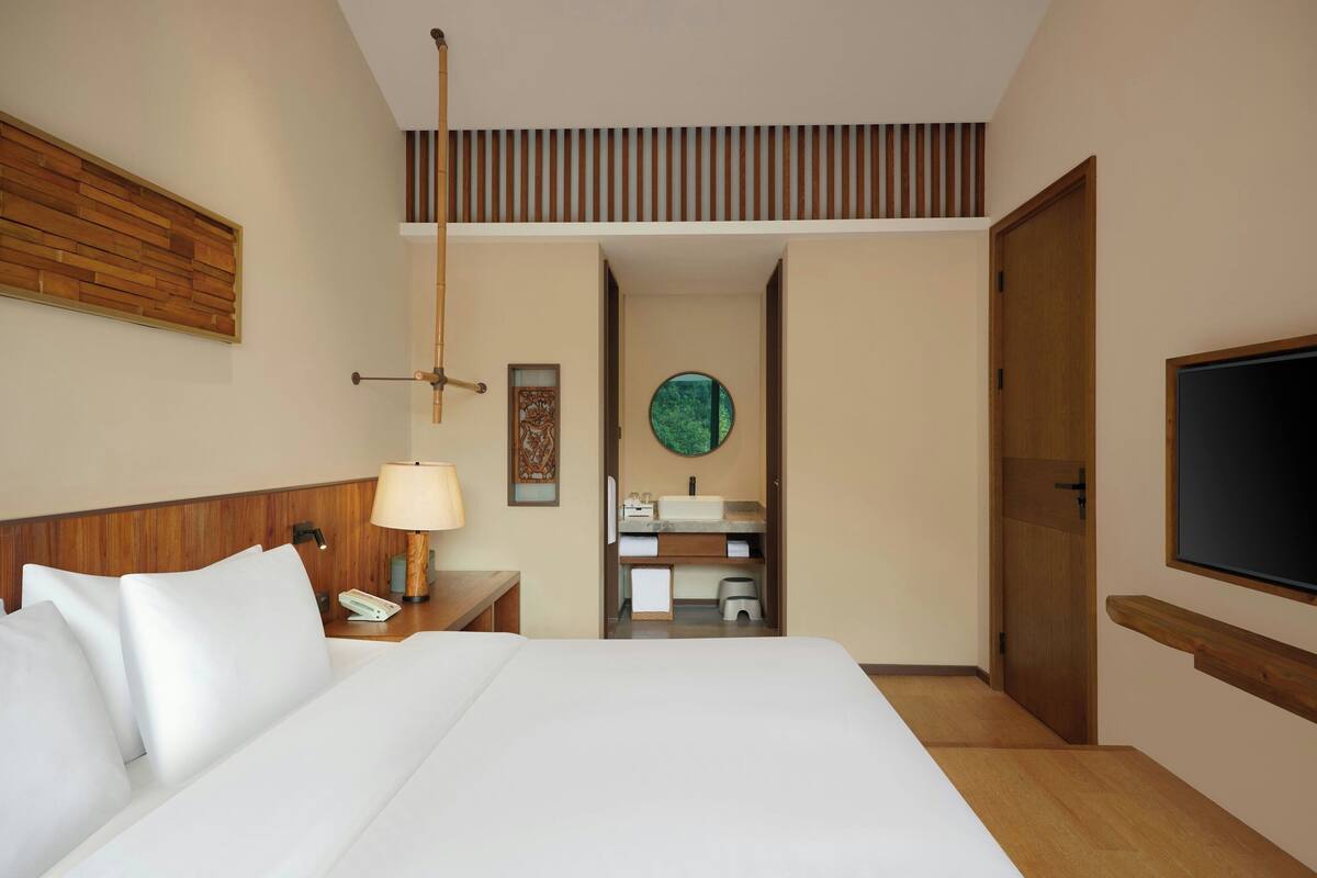 A hotel room with a large bed, a bedside table with a lamp, a TV mounted on the wall, and a wooden headboard.