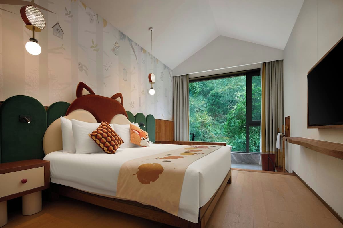 A bedroom with a large bed, a nightstand, a TV, and a window with a view of trees.