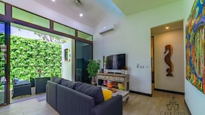 A modern living room with a grey sofa, a flat-screen TV, and a large window overlooking a green garden. - Surf In Surf Out Casa Otto Beachside Colorada (Puntarenas Province)