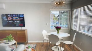 Un salon moderne doté d’un téléviseur à écran plat diffusant un film à suspense, d’un coin repas avec une table ronde et des chaises, et d’une fenêtre équipée de stores.