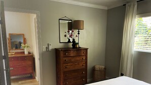 Une chambre à coucher avec une commode en bois, un miroir, une lampe et un lit recouvert d’une couette blanche.