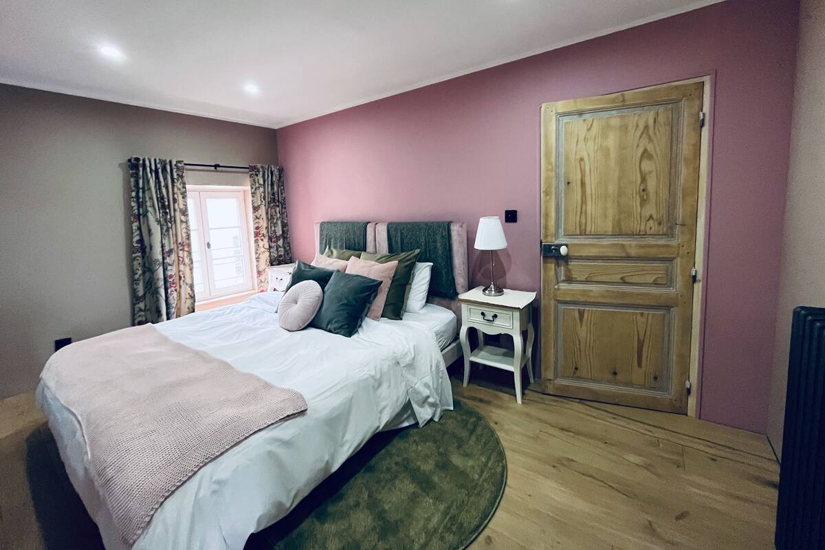 Quarto com cama, mesa de cabeceira, abajur, porta de madeira e janela com cortinas.