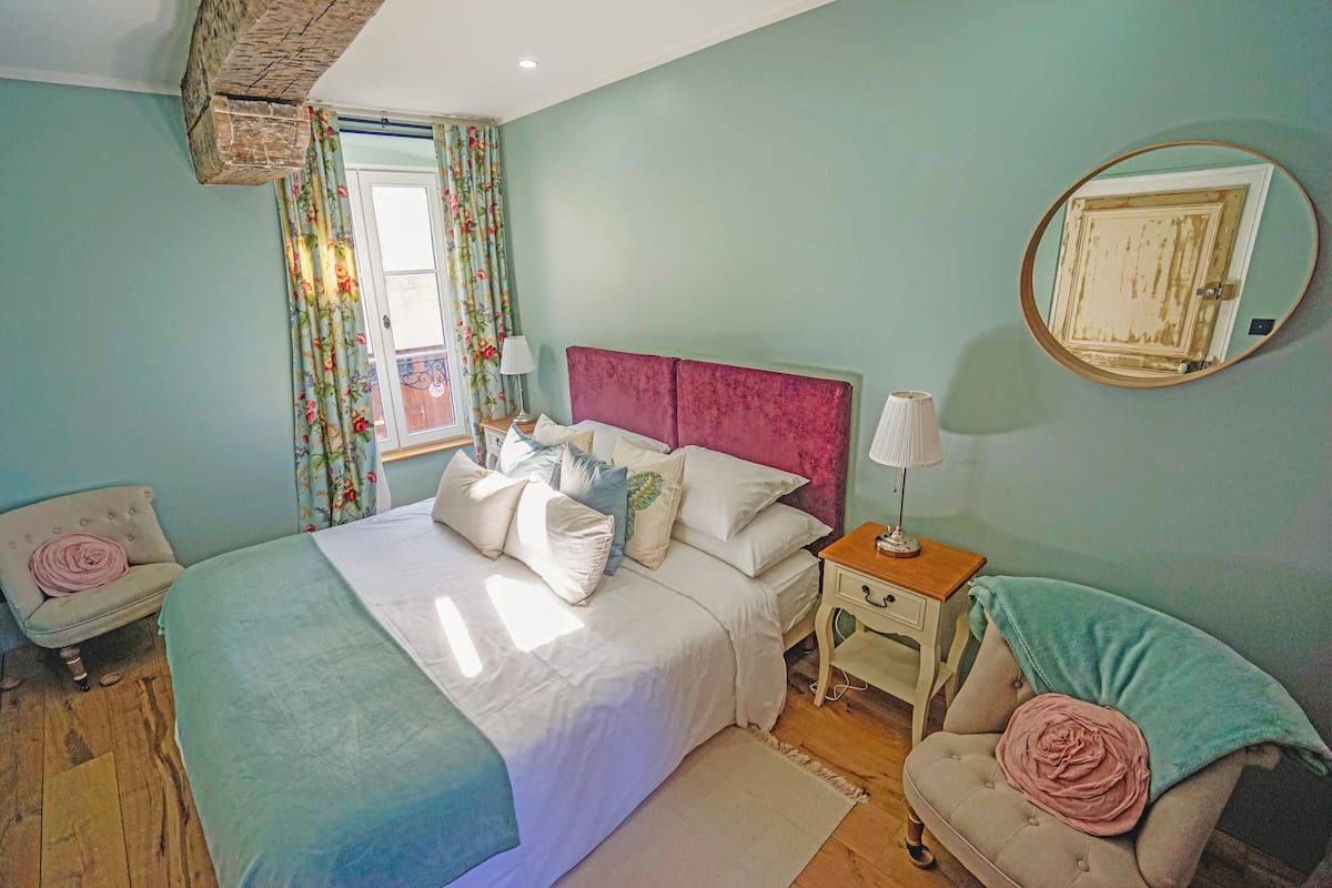 Um quarto com cama, mesas de cabeceira, uma cadeira, um espelho e uma janela com cortinas.