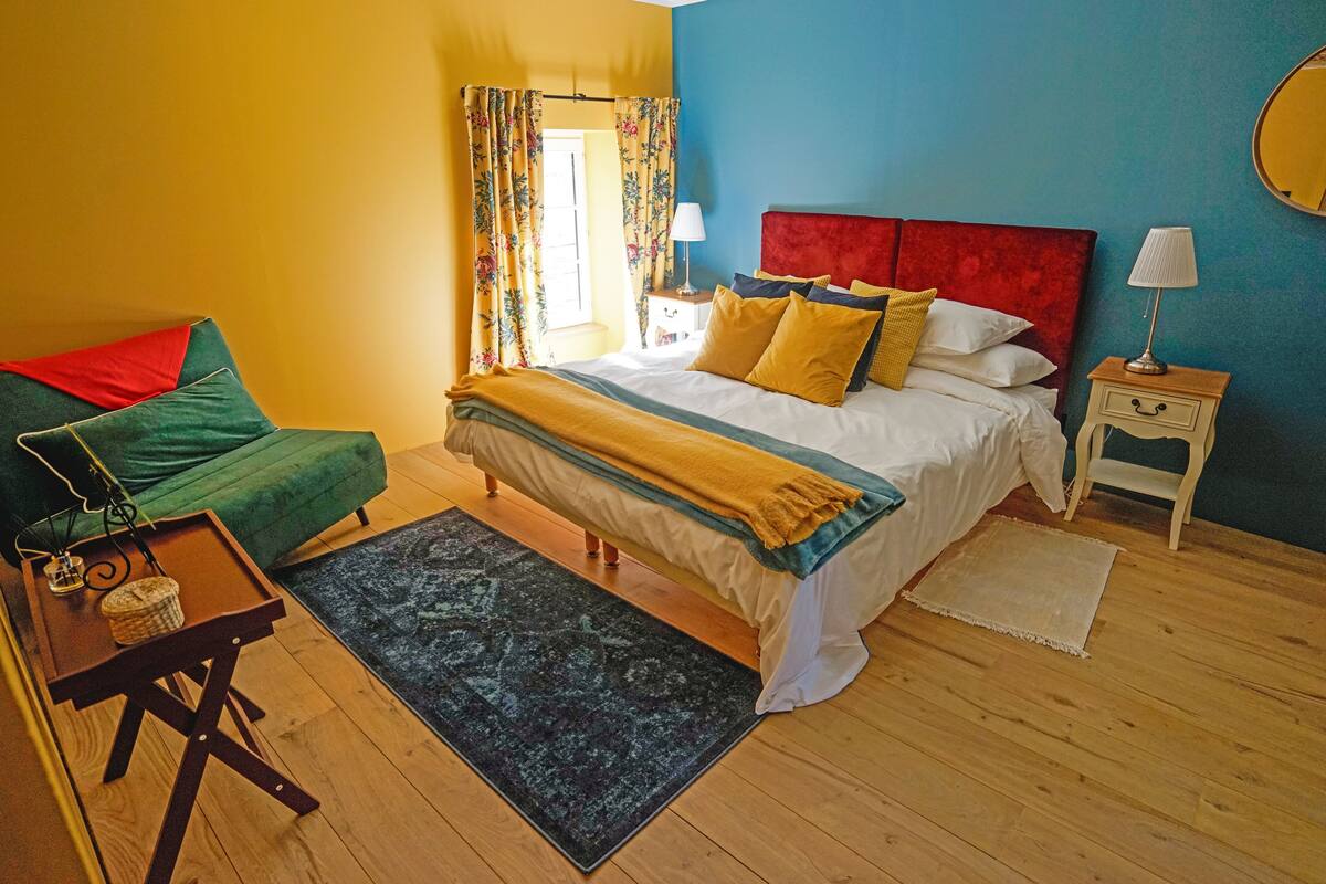 Quarto com cama, mesa de cabeceira, abajur e uma cadeira verde.
