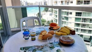 A balcony with a table set for breakfast, including bread, fruits, and beverages, overlooking a cityscape and beach. - Oceanview Apartment in Guarujá: Fast WiFi & Smart TV (Guarujá - SP)
