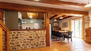 Un muro de piedra, vigas de madera, una cocina, una zona de comedor con sillas y una chimenea de piedra.