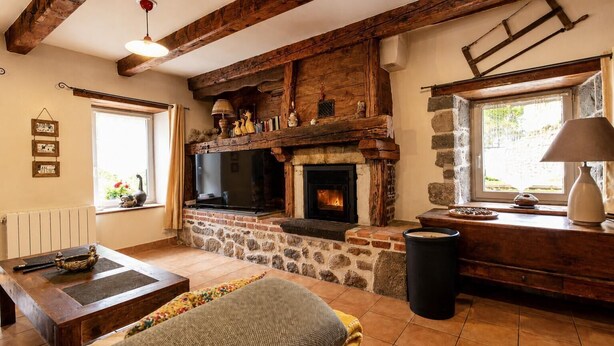 Un interior rústico con vigas de madera a la vista, chimenea de piedra, televisor y ventana con vistas.