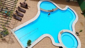 En pool med en ovanlig form, där en person badar, omgiven av solstolar och en pergola.