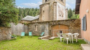 Une cour bordée de murs en pierre, avec une porte en bois et des sièges installés à l’extérieur.