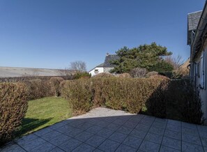 A stone patio leading to a white house with a stone roof, surrounded by hedges and a grassy area.