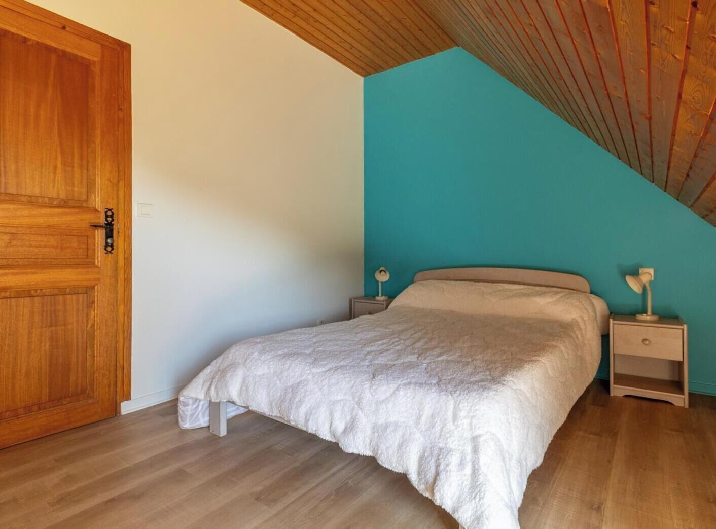 Un dormitorio con puerta de madera, una cama con manta blanca, dos mesitas de noche con lámparas y un techo inclinado.