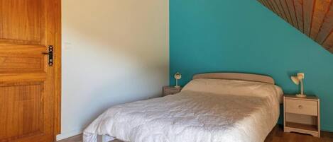 Un dormitorio con puerta de madera, una cama con manta blanca, dos mesitas de noche con lámparas y un techo inclinado.