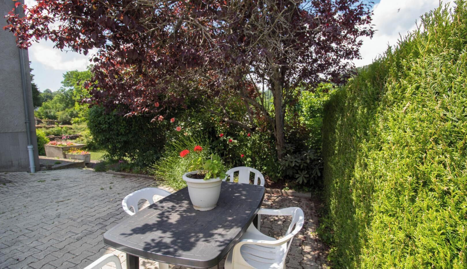 Une terrasse extérieure avec une table et des chaises, entourée de verdure et d’une haie.
