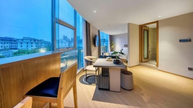 A modern hotel room with a large window, a wooden bench, a small table with a plant, and a view of the cityscape. - Hotel Jagri Taizhou (Taizhou)