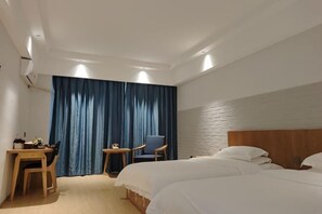 A hotel room with two beds, a desk with a chair, a small table, and a large window with blue curtains.