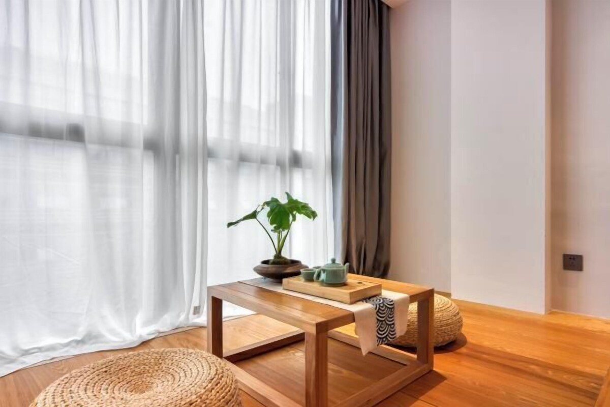 A room with a wooden floor, a small table with a plant and teapot, a woven pouf, and sheer curtains.