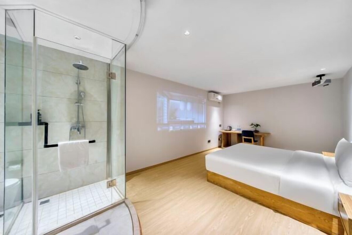 A modern hotel room with a glass-enclosed shower, a bed with a wooden headboard, a small desk, and a projector screen on the wall.