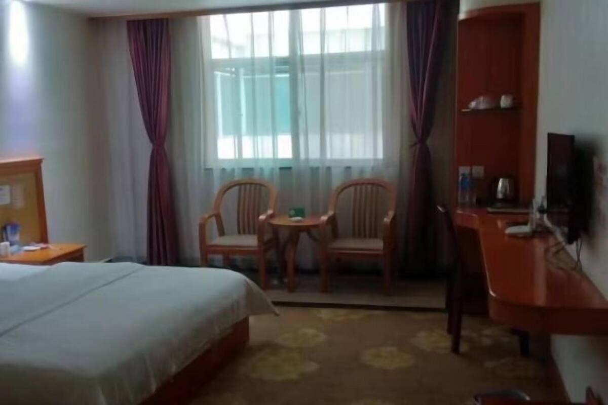 A hotel room with a bed, two chairs, a small table, a desk, a TV, and a window with curtains.