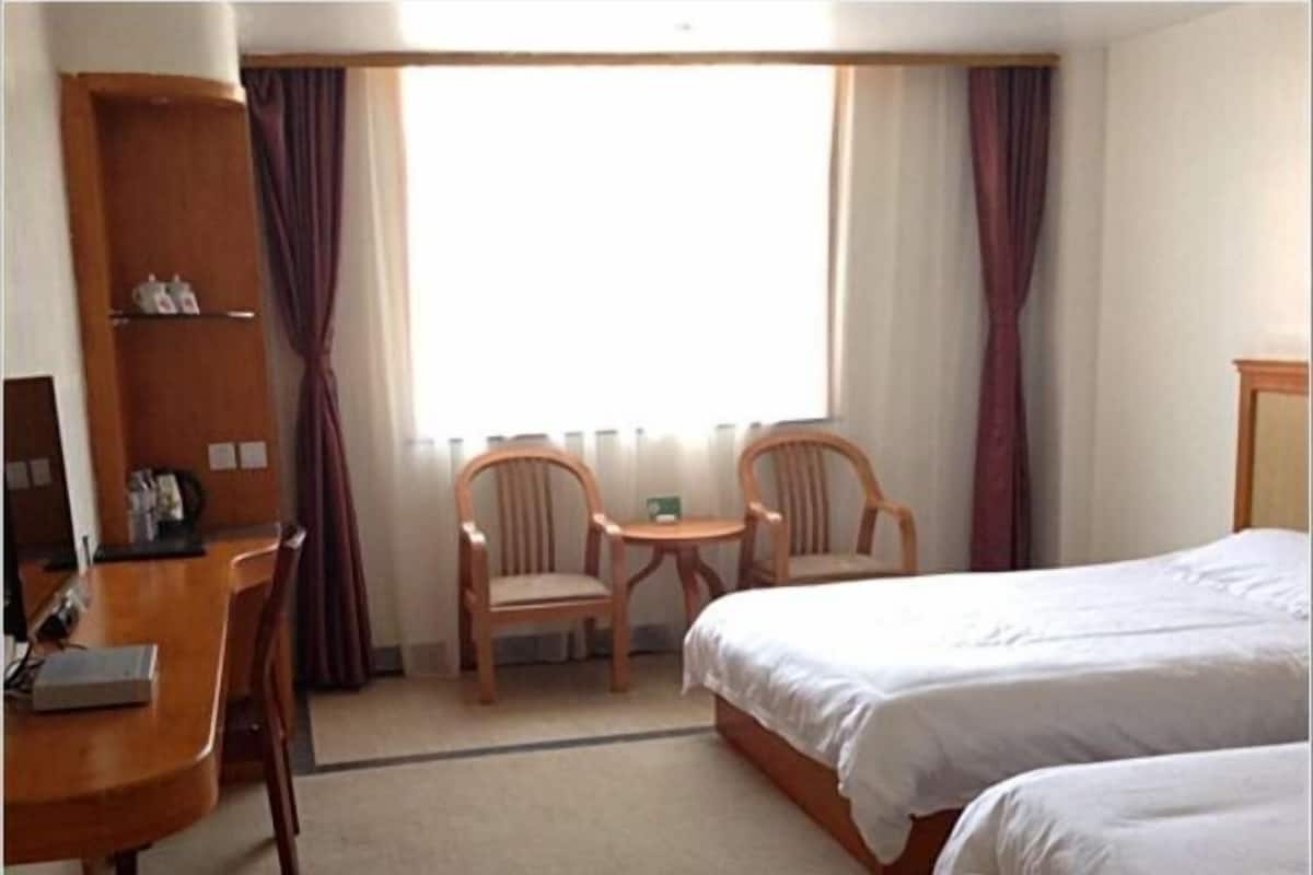 A hotel room with a bed, two chairs, a small table, a wooden shelf, and a window with curtains.