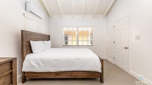 Ein Schlafzimmer mit einem hölzernen Bett, weißer Bettwäsche, einem Fenster mit Jalousien, einer Klimaanlage und einer weißen Tür.