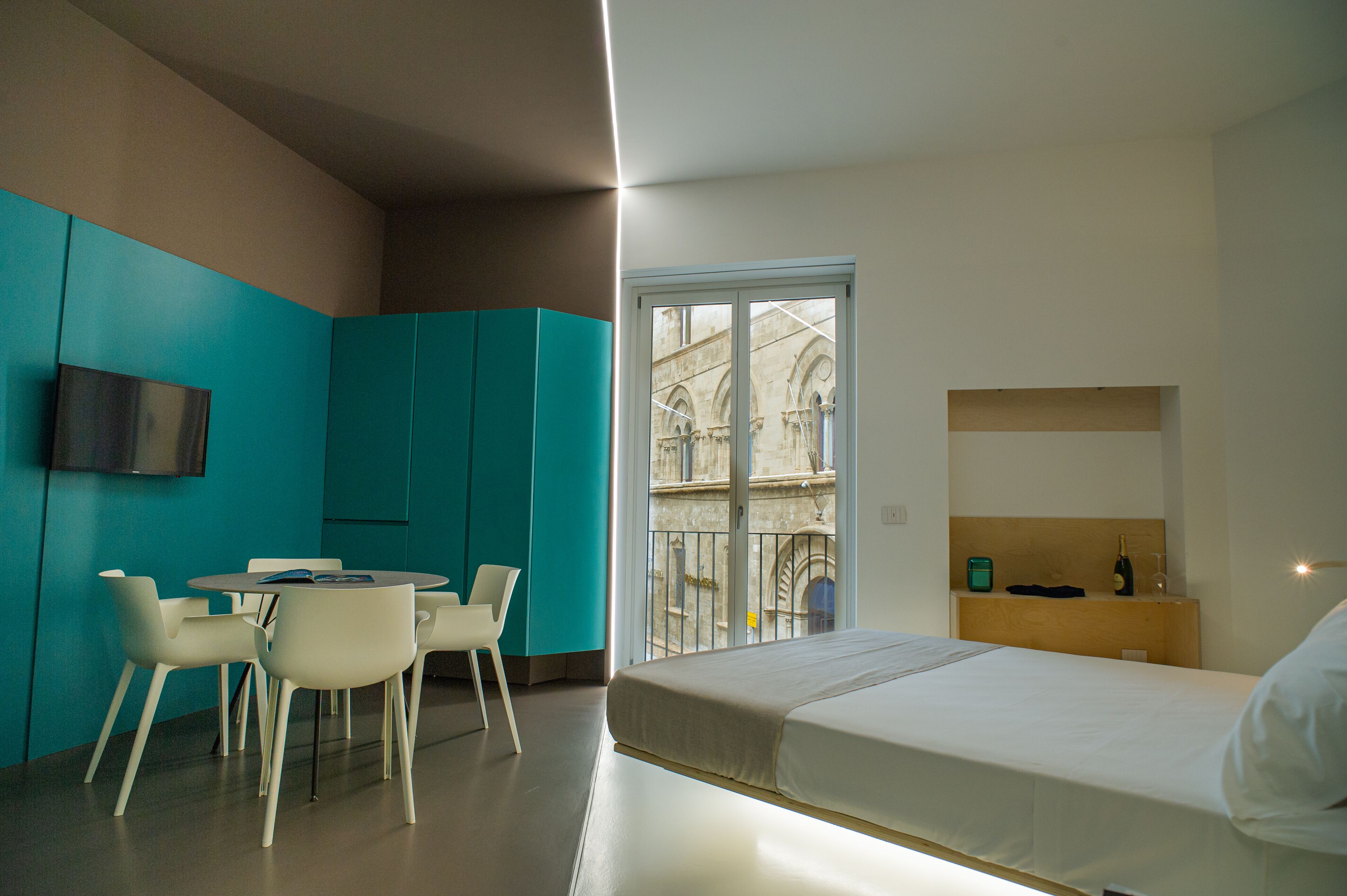 Une chambre d’hôtel moderne équipée d’un lit, d’une petite table avec des chaises, d’un téléviseur fixé au mur et d’une fenêtre donnant sur un bâtiment historique.