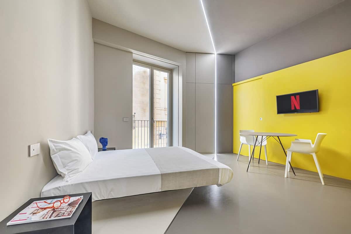 A modern hotel room with a bed, a small table with a magazine, a TV on the wall, and a bright yellow accent wall.