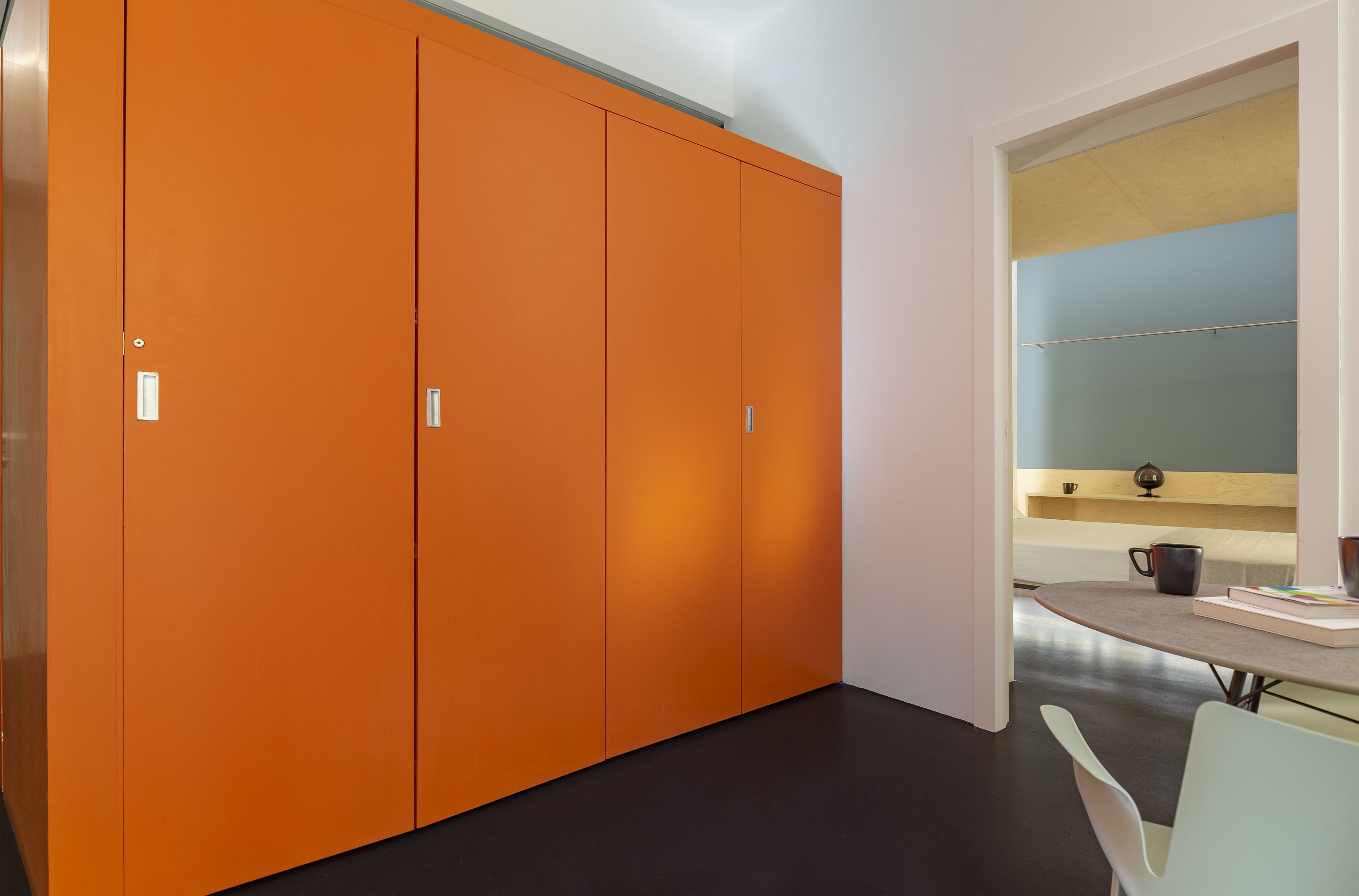 A room with a large orange wardrobe, a small table with a cup and book, and a view of a blue wall and a shelf with a decorative item.
