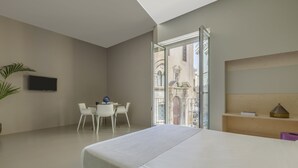 A modern hotel room with a large bed, a desk with chairs, a TV, and a balcony with a view of buildings. - Fiveplace Design Suite & Apartments (Trapani)